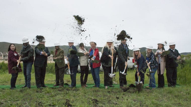 thirteen people in white hard hats holding shovels fling dirt into the air as a celebration of breaking ground at the new facility site